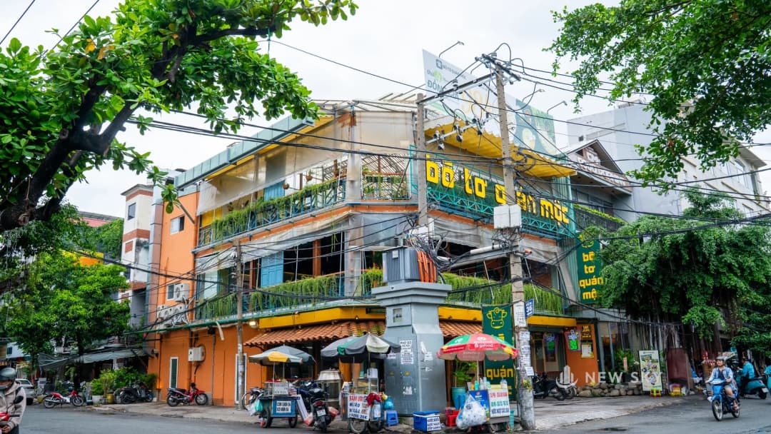 Nhà hàng Bò Tơ Quán Mộc ở góc phố nhìn từ xa: hàng chữ nổi vàng chạy ngang tầng trên, một cột điện bê tông và tủ điện xám dựng ngay trước hàng chữ, chằng chịt dây điện vắt ngang mặt tiền, cành cây lớn xòa xuống từ mép trái ảnh.