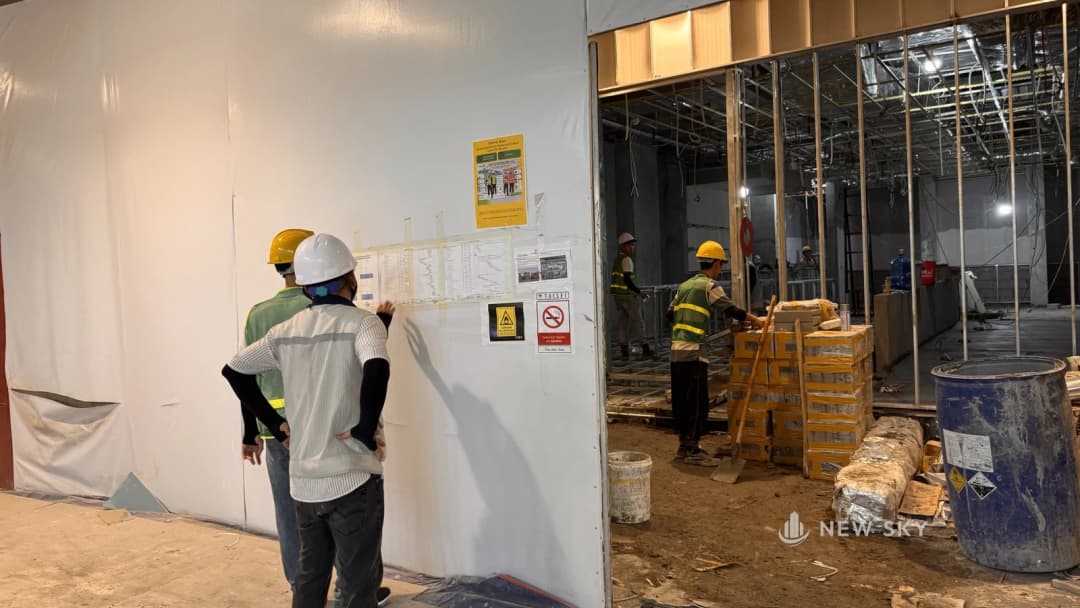 Hai người đội mũ bảo hộ đứng xem hồ sơ dán trên vách quây một gian hàng trong trung tâm thương mại, bên trong thợ làm dưới sàn giữa các chồng vật liệu, khung trần đã dựng xong.