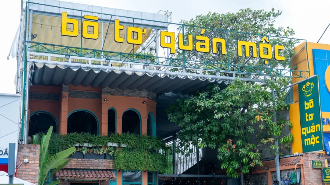 Mặt tiền nhà hàng Bò Tơ Quán Mộc, chữ nổi vàng cỡ lớn gắn trên khung thép cao, tán cây xanh bên phải trùm lên khung và tấm biển dọc ở góc.