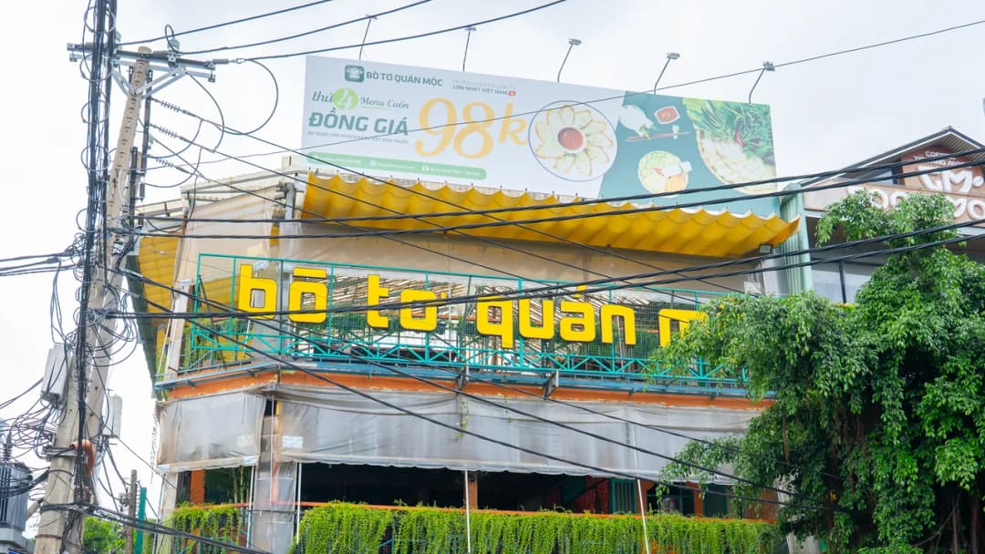 Mặt tiền nhà hàng nhìn từ ngoài đường: bảng quảng cáo khổ lớn dựng trên mái, dưới đó là hàng chữ nổi tên nhà hàng bị tán cây bên phải che mất chữ cuối, phía trước là lưới dây điện chằng chịt.
