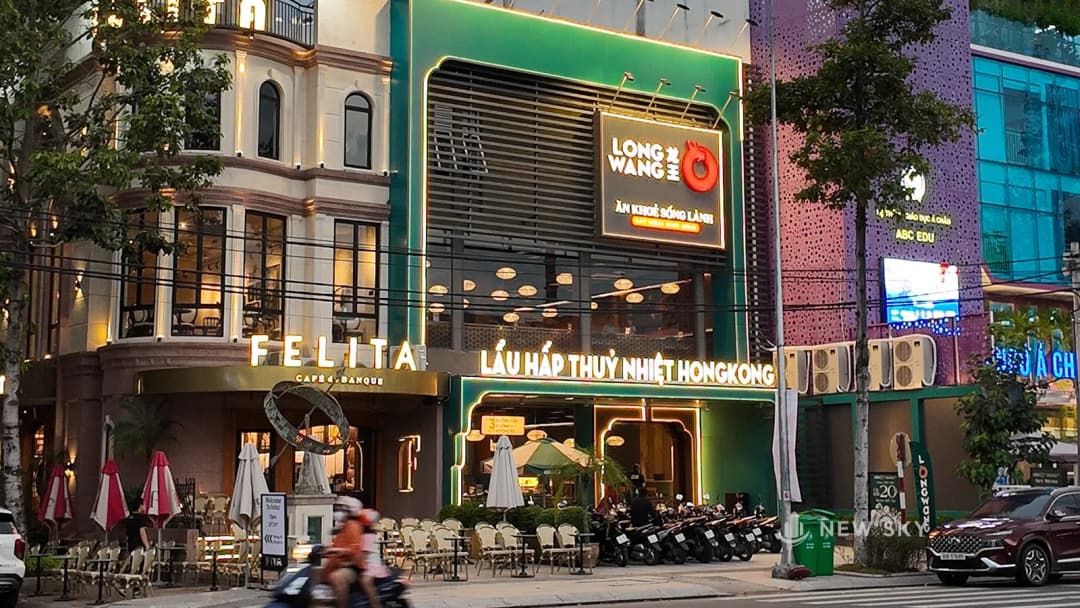 Mặt tiền nhà hàng Long Wang lúc chạng vạng: khối nhà sơn xanh lá, biển logo đỏ trên cao, dải chữ tên món chạy ngang mặt tiền, đèn viền đã bật.