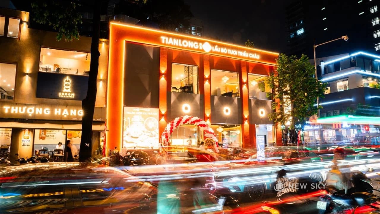 Mặt tiền nhà hàng Tian Long lên đèn buổi tối: khung biển hiệu chữ nổi phát sáng màu cam chạy suốt hai tầng mặt kính, cổng bóng bay đỏ trắng trước lối vào, vệt đèn xe kéo dài trên mặt đường phía trước.