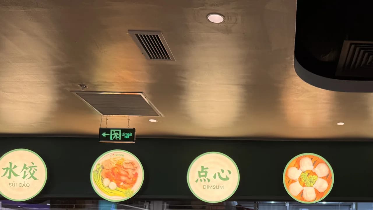 Trần phía trên quầy bếp mở của Cảo Vương Vincom Mega Mall Ocean Park: hai miệng gió dạng lá sách trên trần sơn tối, biển lối thoát hiểm treo giữa, bên dưới là dãy hộp đèn tròn in chữ sủi cảo, dimsum và hình món ăn.