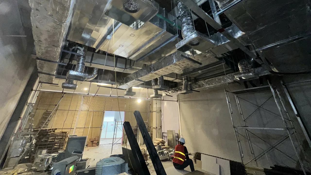 Trần G.Master Aeon Mall Hạ Long lúc đang thi công: ống gió tôn bọc bảo ôn, nhiều nhánh ống mềm thả xuống các hộp miệng gió; một thợ đội mũ bảo hộ ngồi quay lưng ở góc dưới, cạnh giàn giáo.