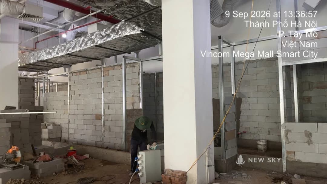 Một mặt bằng đang ở chặng thô tại Vincom Mega Mall Smart City: tường gạch bê tông nhẹ đã dựng gần tới trần, khung thép vách dựng sẵn, phía trên là ống gió bọc bảo ôn và ống nước chữa cháy sơn đỏ còn lộ ra vì trần chưa đóng; một thợ đội mũ vải cúi xuống bên một tủ điện thi công tạm đặt giữa sàn.