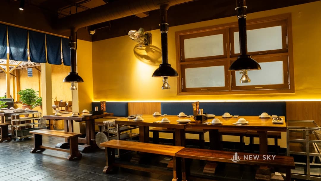 Phòng ăn một cơ sở Khèn Nướng Sapa: ba chụp hút khói hình chuông màu sẫm treo trên dãy bàn gỗ dài, từ mỗi chụp có một ống đứng thẳng lên trần, phía trên là ống gió tròn chạy ngang trần.