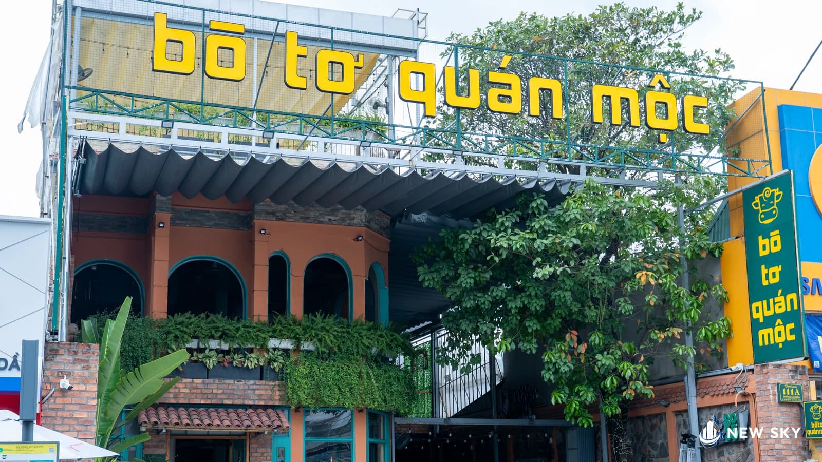 Mặt tiền nhà hàng Bò Tơ Quán Mộc trên nhà phố: biển hiệu chữ vàng cỡ lớn dựng trên khung sắt xanh chạy suốt mái, mái hiên xếp sóng màu than, ban công gạch đỏ phủ cây leo, một cây lớn che bớt lối vào bên phải.