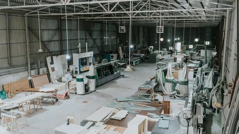 Xưởng nội thất nhìn từ trên cao: máy dán cạnh nằm dọc giữa xưởng, bộ hút bụi túi vải đứng cạnh, tủ trắng ráp dở và ván xếp quanh.