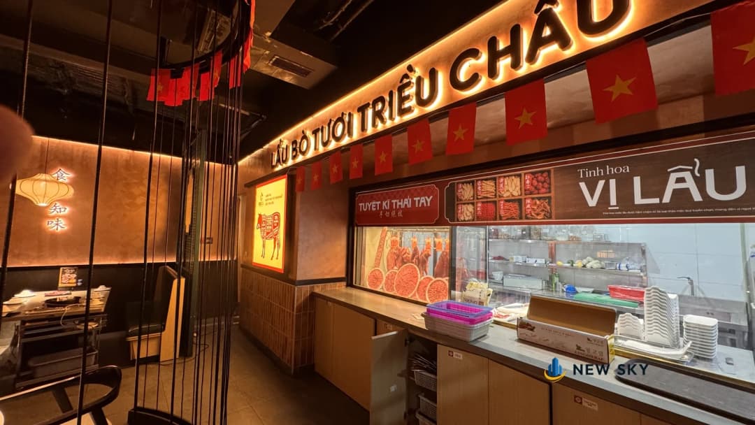 Một dãy quầy chạy dọc tường trong nhà hàng: thân quầy ốp ván vân gỗ sáng màu, mặt quầy sẫm màu, bên dưới là dãy cánh tủ với một cánh đang mở ra. Phía trên quầy là tủ kính trưng bày thịt và biển chữ nổi Lẩu Bò Tươi Triều Châu, cờ đỏ sao vàng treo thành dây dọc trần.