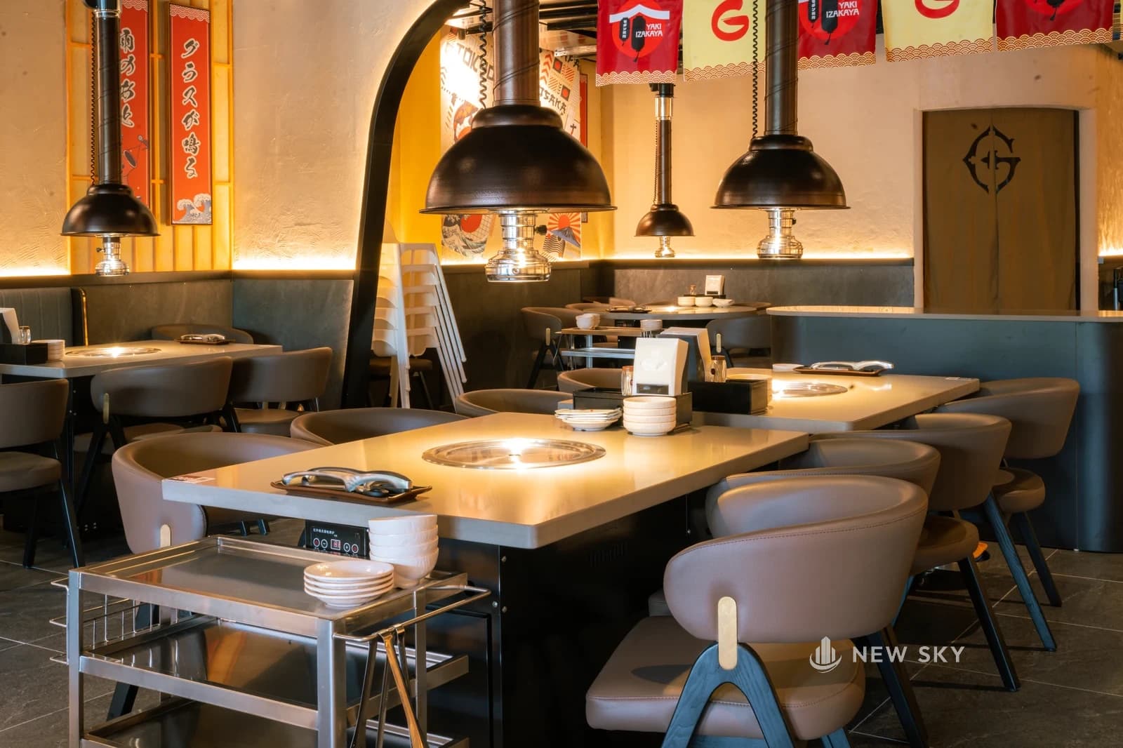 Bàn nướng bốn chỗ mặt đá sáng: bếp âm tròn có vỉ giữa mặt bàn, bảng điều khiển gắn dưới mép bàn, chụp hút hình chuông màu sẫm thả từ trần, xe đẩy inox để bát đĩa đặt sát bàn, phía sau là cửa vòm và dây cờ vải.