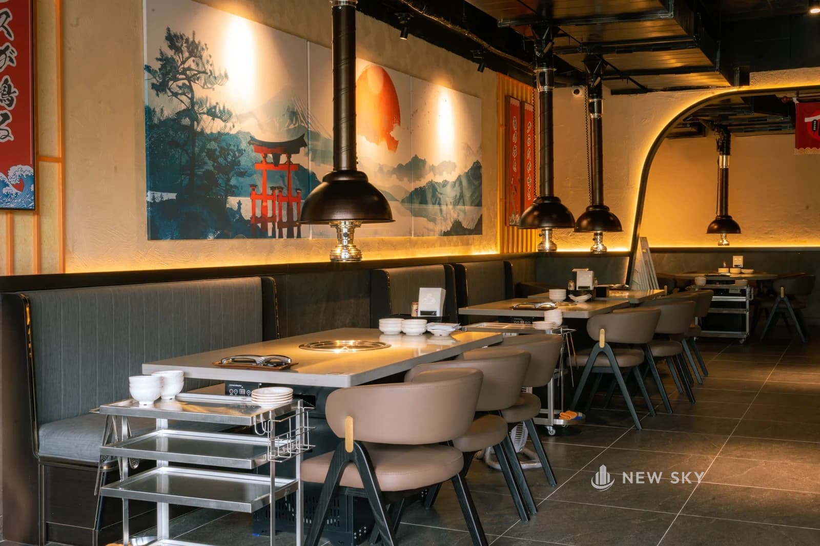 Dãy bàn nướng dựa ghế băng xám dưới bức tranh núi tuyết và cổng torii đỏ, chụp hút hình chuông treo trên từng bếp âm tròn, xe đẩy inox đặt đầu bàn, cổng vòm dẫn sang phòng trong.