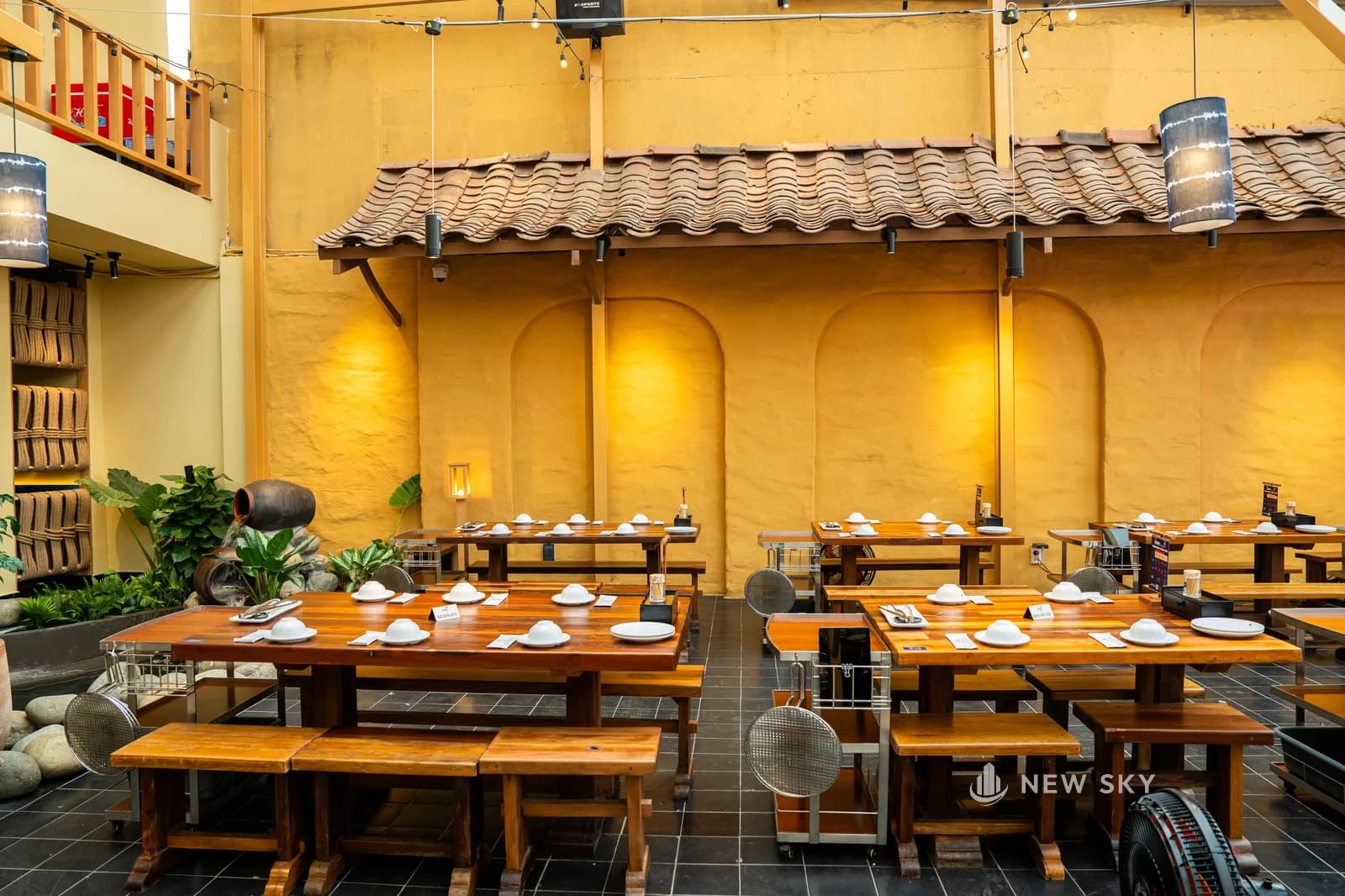 Sân nhà hàng nướng: nhiều bộ bàn gỗ dài kê ghế gỗ không tựa, tường trát vàng khoét hốc vòm, mái ngói gắn dọc tường, đèn lồng vải treo cao, không bàn nào có chụp hút.