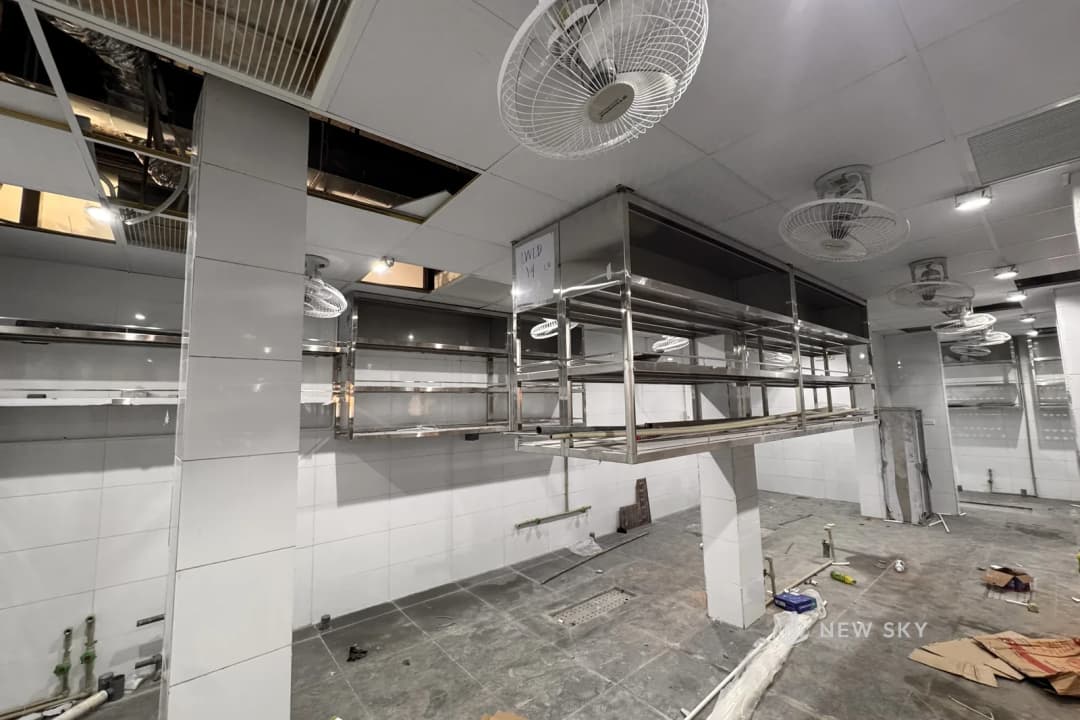 Khu bếp đang thi công: tường ốp gạch men trắng, giá kệ inox nhiều tầng treo trên cao và đứng dưới sàn, ống hút khói inox chạy dọc trần, quạt treo tường, trần còn hở nhiều ô lộ đường kỹ thuật, ống nước chờ nhô lên ở chân tường.