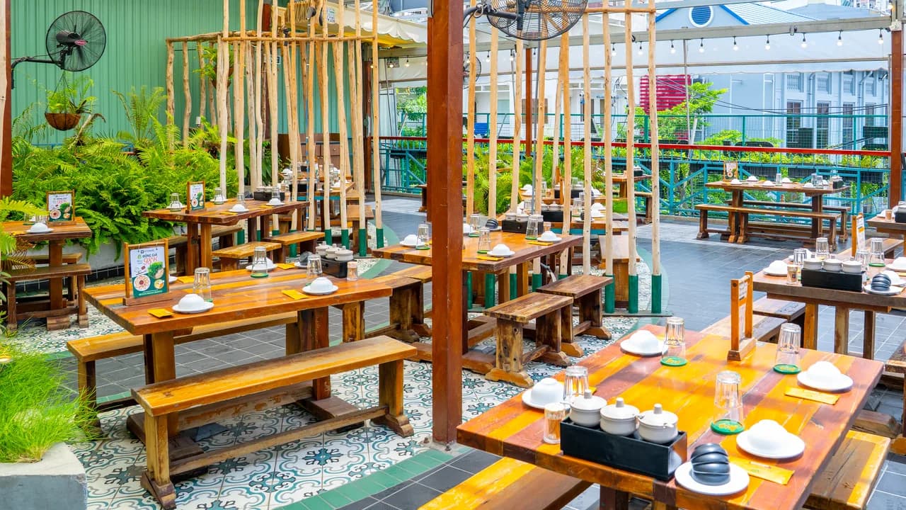 Sân thượng Bò Tơ Quán Mộc ở Võ Oanh: bàn ghế gỗ dài kê trên sàn gạch bông, những thân tre dựng đứng chia khu, chậu dương xỉ dọc tường tôn xanh, quạt treo trên cột, phía xa là lan can và dãy nhà bên kia phố.