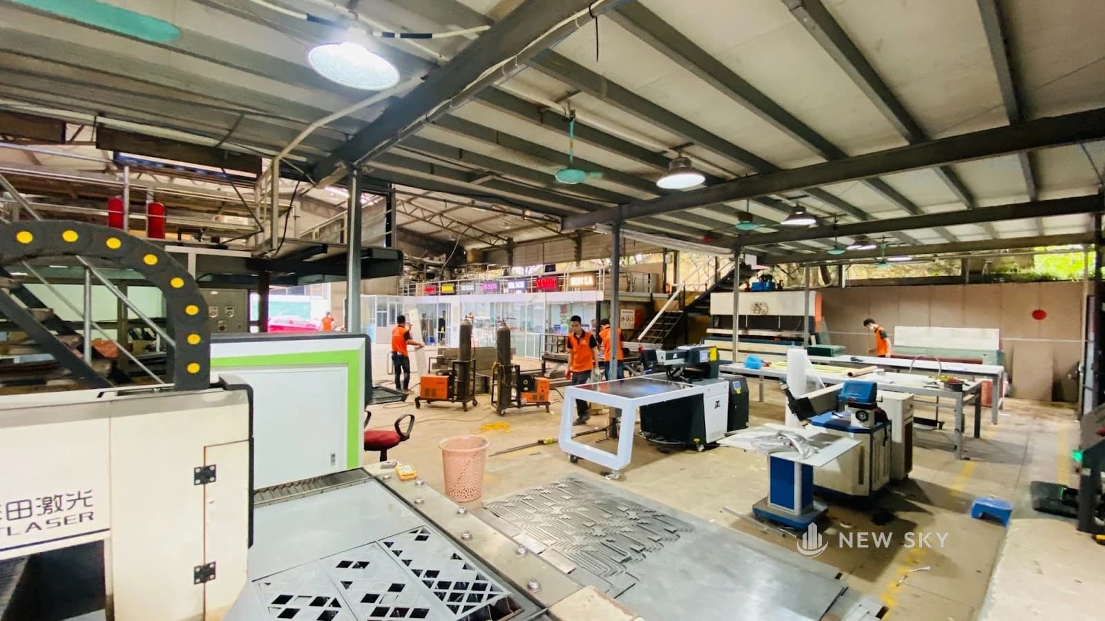 Khu làm quảng cáo ở xưởng Thượng Hồng: máy cắt laser ở tiền cảnh, tấm kim loại vừa cắt hoa văn nằm dưới sàn, thợ mặc áo cam làm ở các bàn phía sau