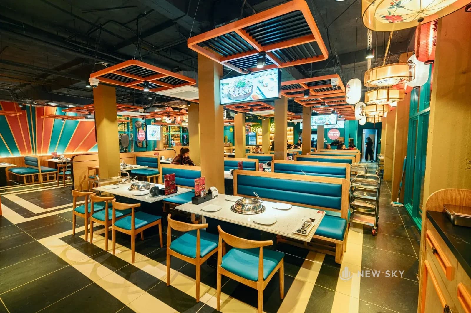 Phòng ăn nhà hàng Long Wang 84 Ngọc Khánh: khoang ghế băng bọc da xanh ngọc, ghế gỗ đệm xanh, bàn mặt đá sáng có nồi inox; dưới trần để hở sơn đen treo những khung lam gỗ sơn cam, màn hình, đèn lồng, máy điều hòa gắn trần và ống gió; bên trái là mảng tường sơn hình quạt tia cam và xanh, sàn gạch sẫm có dải sáng chạy men lối đi.