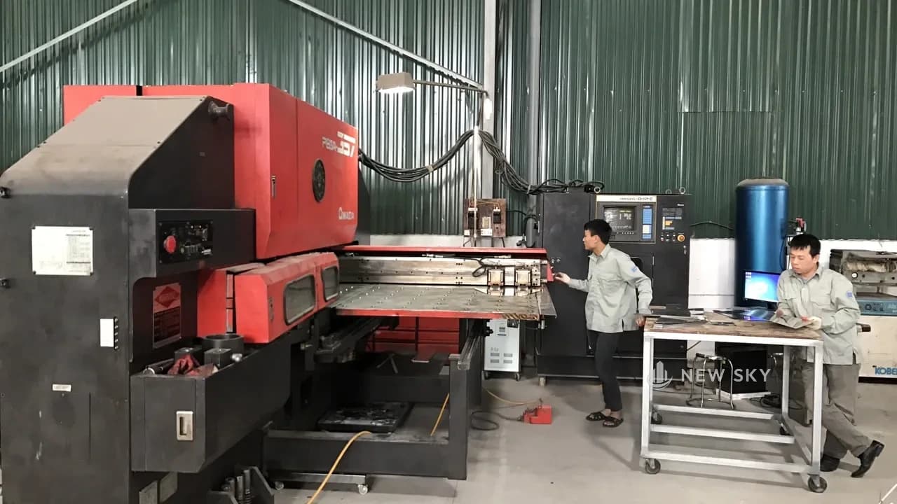 Máy đột dập tấm kim loại trong xưởng New Sky, hai thợ đang căn phôi inox trên bàn máy.