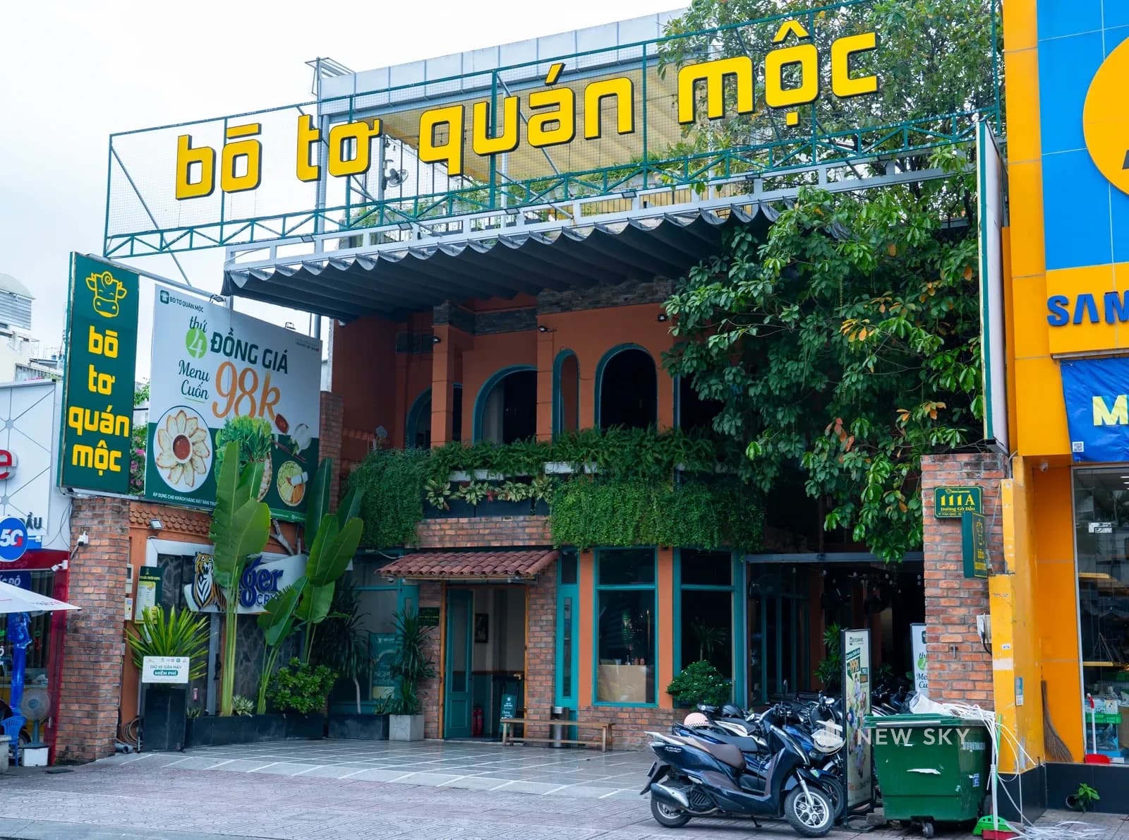 Mặt tiền Bò Tơ Quán Mộc 111 Gò Dầu: nhà hai tầng bo vòm cuốn, dây leo rủ kín ban công, biển chữ vàng gác trên khung sắt chạy suốt bề ngang.