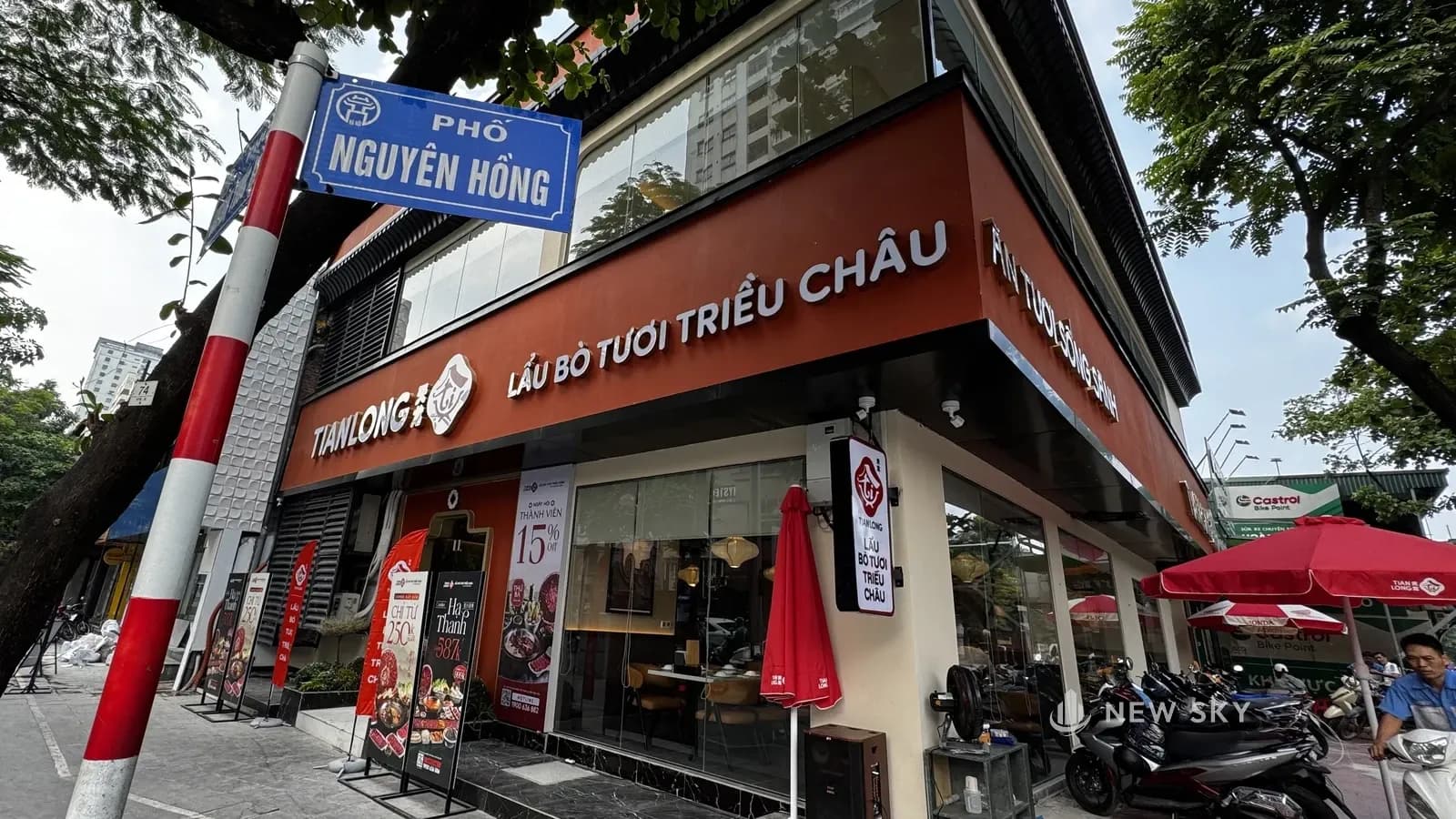 Góc phố Huỳnh Thúc Kháng và Nguyên Hồng: căn nhà 2 tầng của Tian Long, dải biển đỏ cam chạy liền qua hai mặt, mặt này ghi "LẨU BÒ TƯƠI TRIỀU CHÂU", mặt kia ghi "ĂN TƯƠI SỐNG SÀNH", tầng 2 là dải kính quanh góc, biển tên phố Nguyên Hồng ở tiền cảnh.