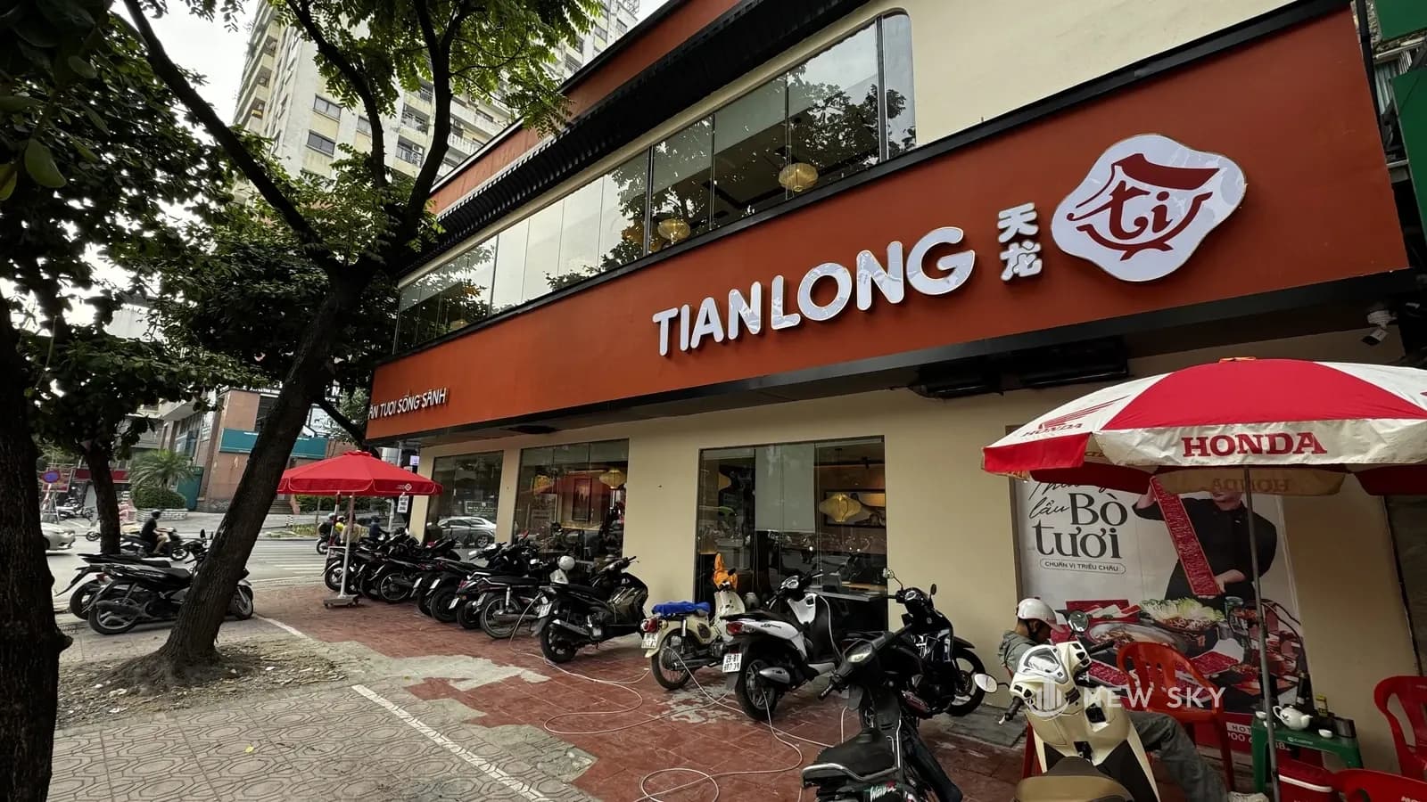 Mặt bên phía phố Nguyên Hồng: dải biển đỏ cam mang chữ "TIANLONG" và logo chạy dọc vỉa hè, tầng 1 là dãy ô kính, xe máy đỗ trước nhà hàng dưới mấy chiếc ô đỏ.
