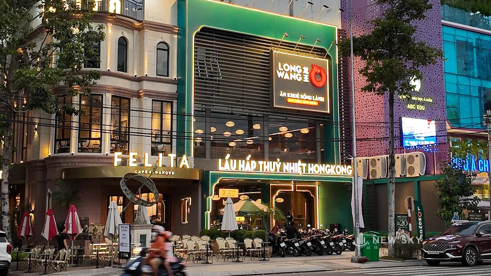Mặt tiền nhà hàng Long Wang lúc chạng vạng: khối nhà sơn xanh lá, biển logo đỏ trên cao, dòng chữ "LẨU HẤP THUỶ NHIỆT HONGKONG" phát sáng chạy ngang tầng trệt.