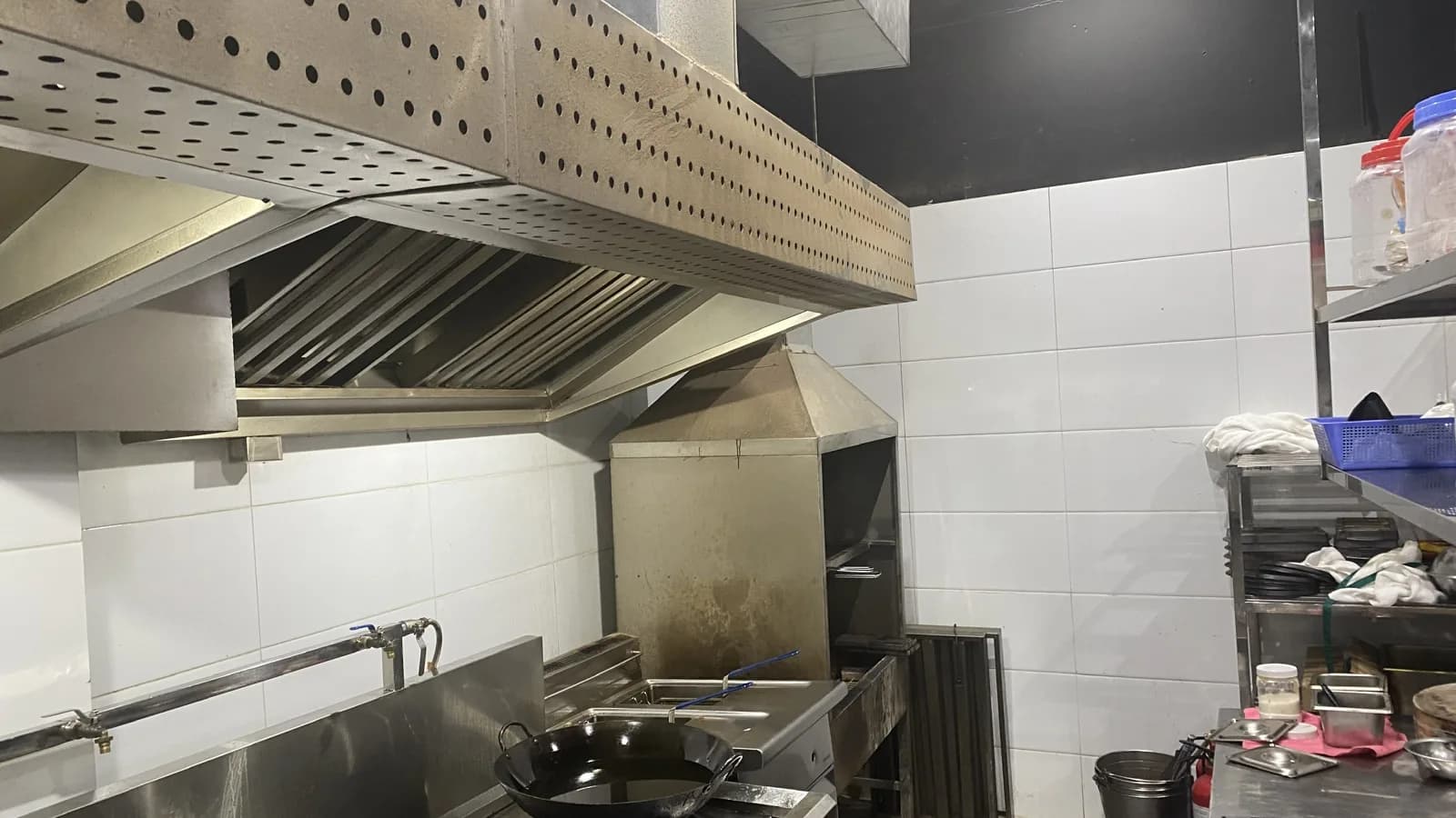 Bếp Bò Tơ Quán Mộc Gò Dầu: chụp hút inox đục lỗ chạy dài trên dãy bếp đặt chảo, bên dưới là hàng lưới lọc mỡ; kế bên dãy bếp là bếp chiên, giá inox và một tấm lọc mỡ dựng sát chân tường.