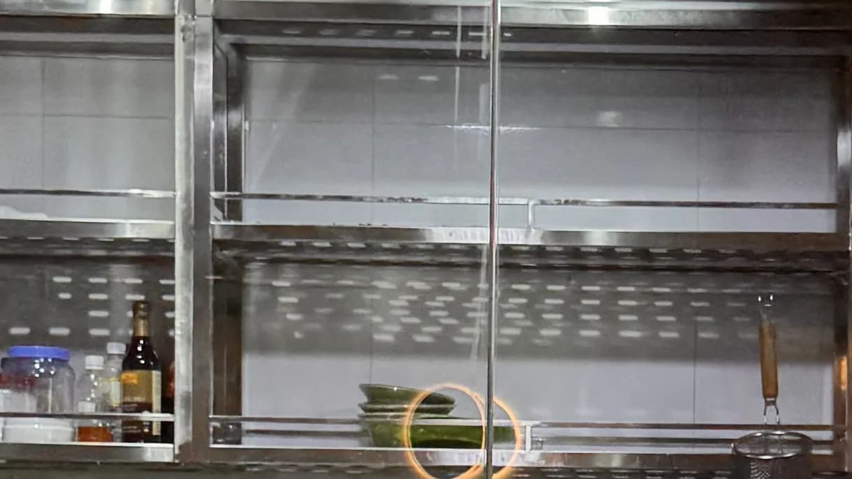 Giá inox treo tường trong bếp mở của Cảo Vương Vincom Mega Mall Ocean Park, chụp qua vách kính quầy: hai tầng giá khung inox, tầng dưới lót tấm đục lỗ, trên giá có chai lọ gia vị, chồng bát và một vợt lọc.