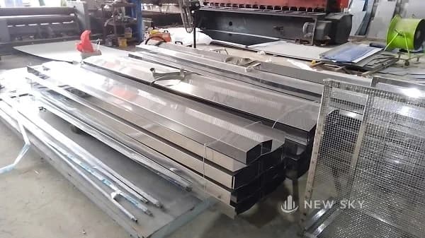 Tấm inox và hộp inox đã chấn xếp thành dãy trên sàn xưởng, phía sau là máy chấn và bàn máy, bên phải là xe đẩy lưới.
