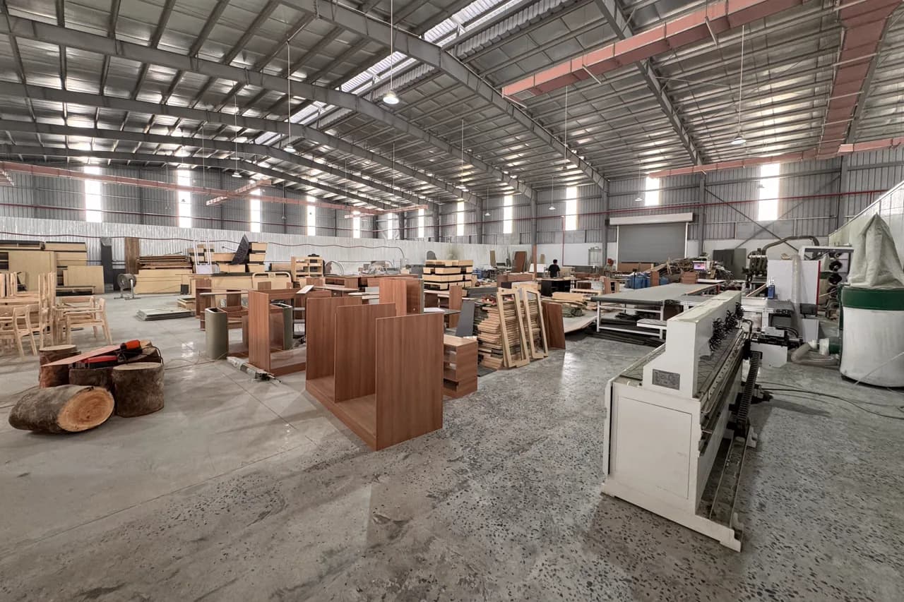 Khu đồ gỗ trong nhà xưởng kèo thép mái cao: thân tủ gỗ màu nâu đang đóng dở xếp giữa sàn, mấy khúc gỗ tròn ở bên trái, máy dán cạnh màu trắng ở tiền cảnh bên phải.
