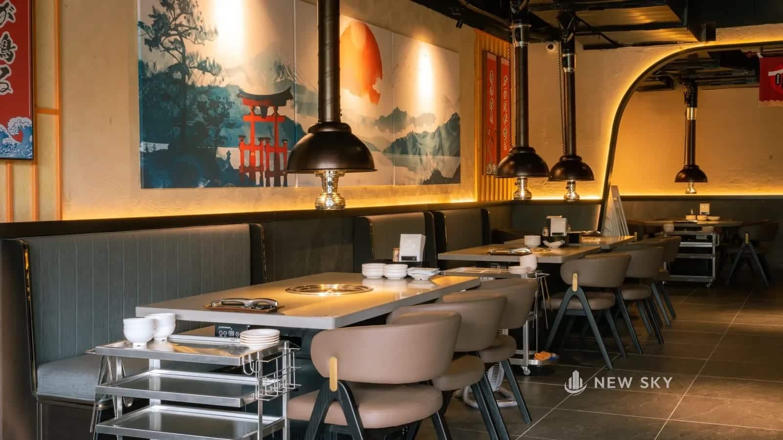 Hai dãy bàn kê song song trong phòng ăn, giữa hai dãy là lối đi, mỗi bàn có bếp âm ở giữa và chụp hút phía trên.