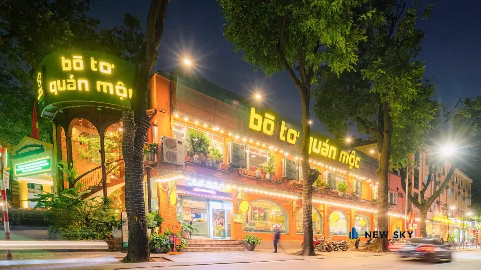 Mặt tiền nhà hàng Bò Tơ Quán Mộc ở số 1 Tăng Bạt Hổ, Hà Nội buổi tối: biển chữ đèn trên khối tháp góc phố và dải chữ vàng chạy dọc mặt phố bên.