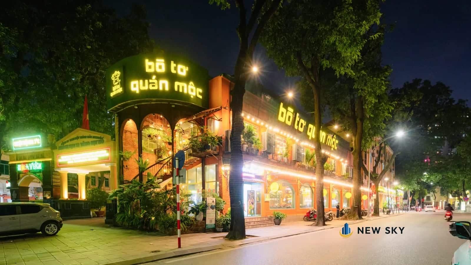 Nhà hàng Bò Tơ Quán Mộc ở góc hai phố tại số 1 Tăng Bạt Hổ, Hà Nội: biển chữ đèn đặt trên khối tháp góc, dải chữ chạy dọc mặt bên, sau hàng cây vỉa hè.