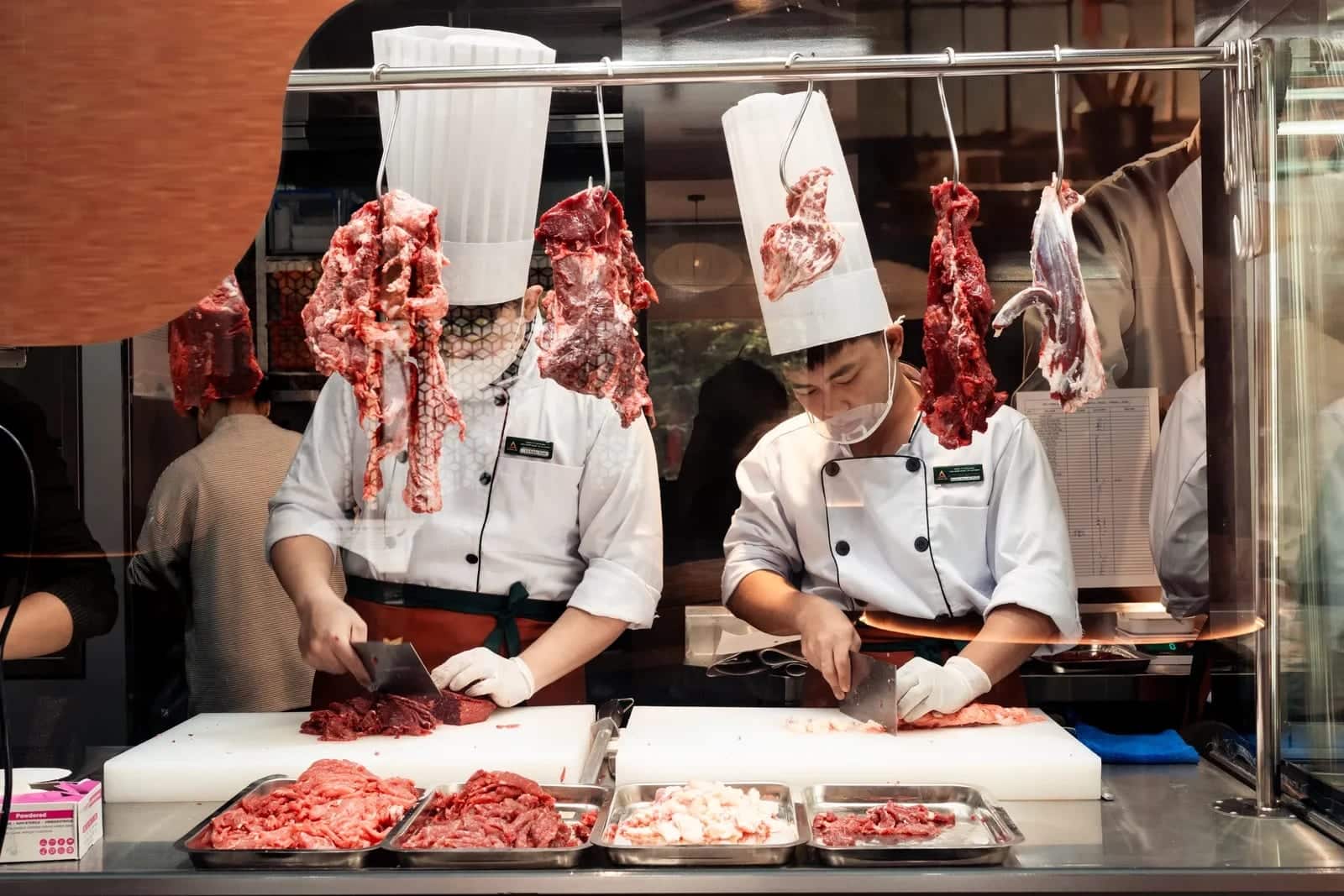 Hai đầu bếp thái thịt sau quầy bếp mở của nhà hàng Tian Long, mặt bàn thao tác và khay đựng đều bằng inox.