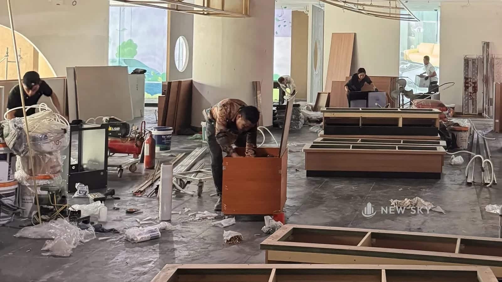 Khu khách nhà hàng Lẩu Ngũ Phúc đang lắp nội thất: thợ ráp tủ gỗ, khung bàn gỗ đặt trên sàn, tấm ván, thùng sơn và dây ống nằm rải quanh.