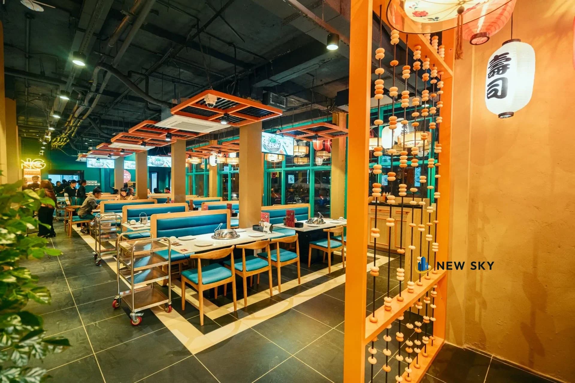 Phòng ăn Long Wang trong tổ hợp 84 Ngọc Khánh nhìn hút vào trong: dãy bàn lẩu chạy dọc, cột vuông phủ màu cam đất, vách rèm hạt gỗ và đèn lồng treo bên phải.