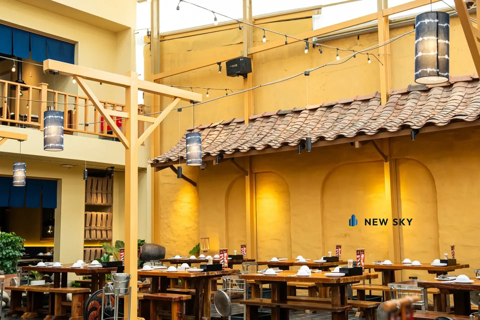 Sân trong nhà hàng Khèn Nướng Sapa số 9 Thoại Ngọc Hầu: bức tường trát tay sơn vàng khoét những hốc vòm âm có đèn rọi, dải mái ngói đỏ nâu lợp chạy dọc tường, đèn lồng hình ống màu chàm treo trên khung sơn vàng, dây bóng đèn chăng ngang, dãy bàn gỗ và ghế dài không tựa bên dưới, ban công lan can gỗ tầng trên ở góc trái.