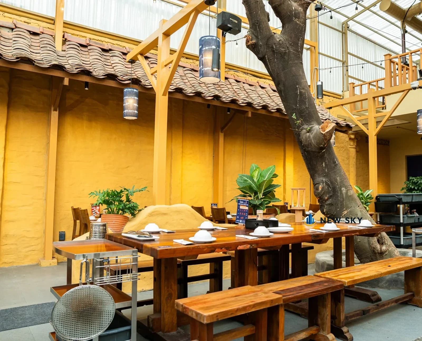 Sân trong Khèn Nướng Sapa 9 Thoại Ngọc Hầu: một thân cây lớn nghiêng ngang qua giữa sân, mái ngói dốc chạy dọc tường trình đất, đèn lồng vải chàm thả dưới khung gỗ, phía trên là mái lấy sáng thông lên tầng lửng.