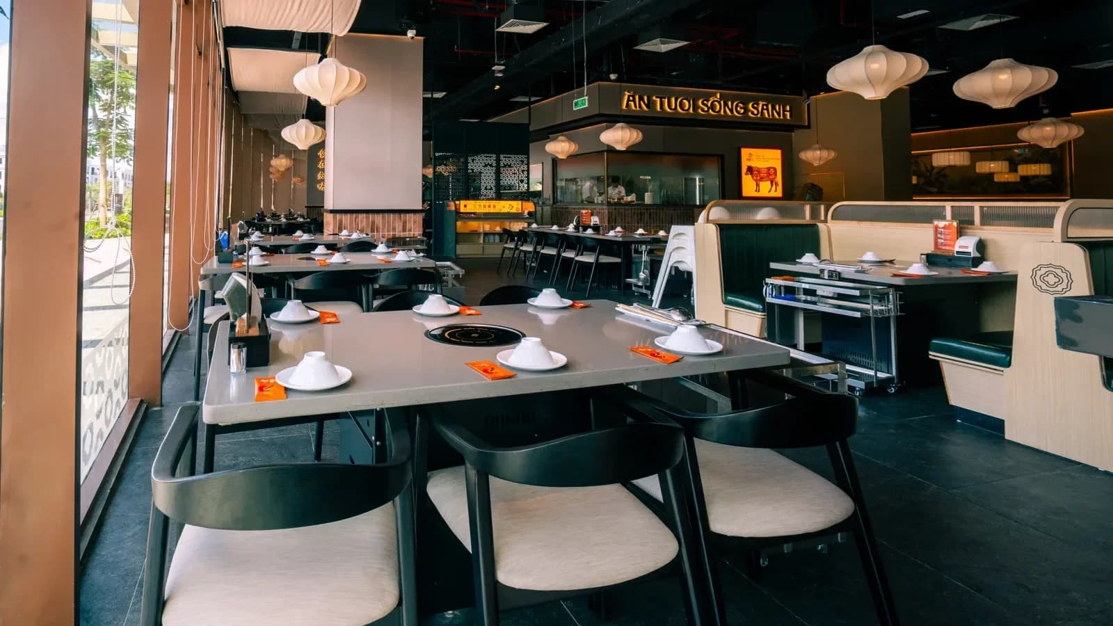 Khoang phòng ăn với trần đen lộ hệ kỹ thuật, nan gỗ, đèn lồng vải và biển chữ sáng phía quầy bếp mở.