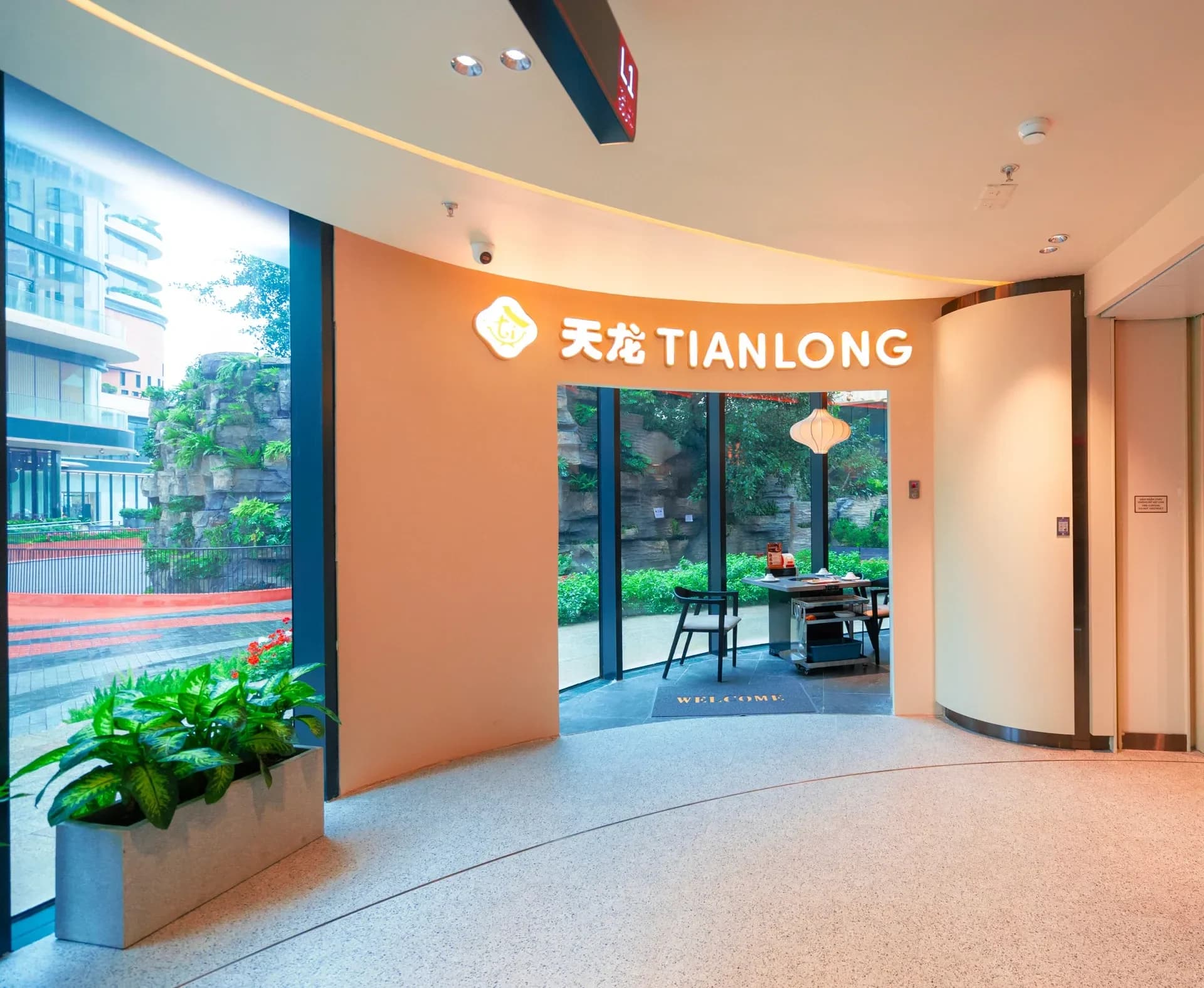 Lối vào nhà hàng Tian Long ở Hà Nội Center: vách cong sơn màu cam đào, chữ 天龙 TIANLONG và hoa Tianlong phát sáng phía trên cửa, qua cửa thấy một bàn lẩu và đèn lồng vải trắng bên vách kính nhìn ra vườn.