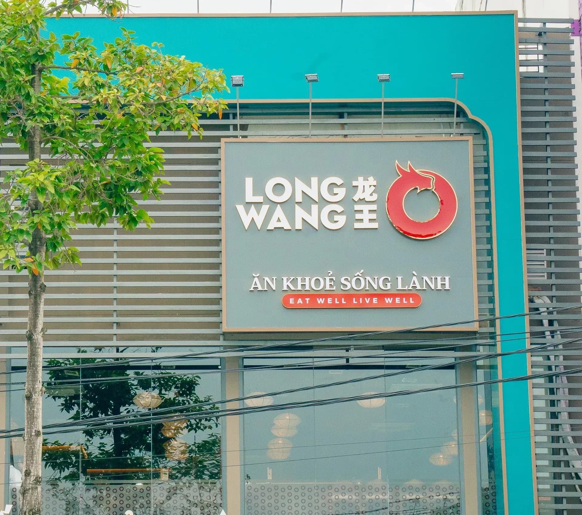 Tầng trên mặt tiền nhà hàng Long Wang ở Biên Hoà, Đồng Nai ban ngày: khung viền xanh ngọc, lam che nắng màu xám, biển LONG WANG 龙王 kèm logo vòng tròn đầu rồng màu đỏ và dòng Ăn khoẻ sống lành, bên dưới là vách kính tầng 2 có đèn lồng tròn, bên trái một cây xanh trước mặt tiền.