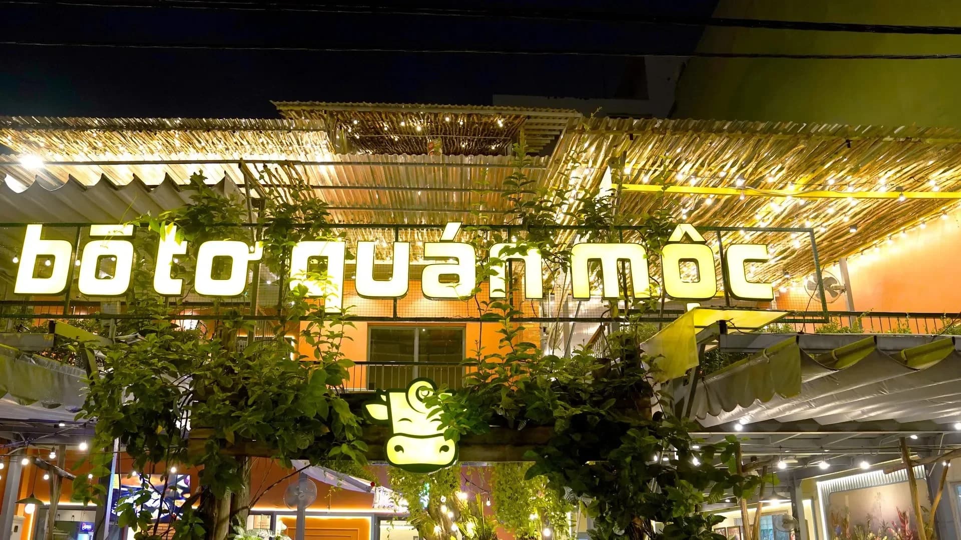 Sân trước nhà hàng Bò Tơ Quán Mộc ở O1 Trường Sơn ban đêm: chữ rời tên nhà hàng phát sáng dựng trên khung trước giàn mái nan tre, dây bóng đèn tròn chăng dưới mái, cây leo phủ khung, logo đầu bò phát sáng trên cổng cây, phía sau là tường nhà sơn cam.