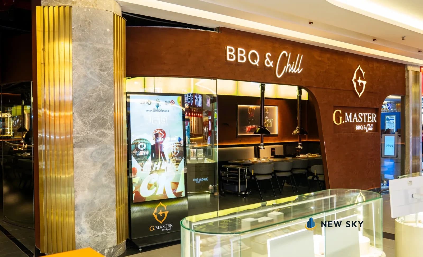 Mặt tiền G.Master BBQ & Chill trong trung tâm thương mại: vách ốp nâu gắn chữ nổi sáng đèn, cột ốp kim loại gân dọc và tủ kính trưng bày cạnh lối vào.