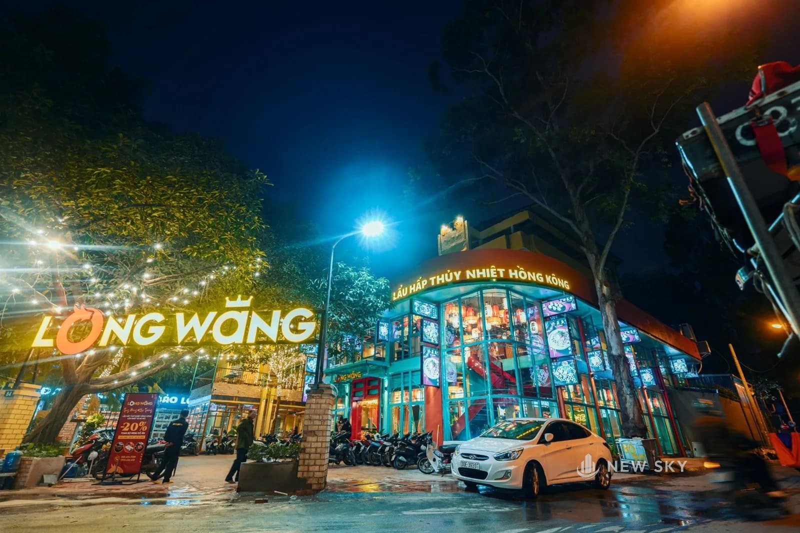 Mặt tiền Long Wang ở tổ hợp 84 Ngọc Khánh buổi tối: khối kính cong hai tầng sáng đèn, biển chữ nổi dựng trên mái khu nhà bên cạnh, đèn dây giăng trên tán cây, xe máy đỗ kín sân.