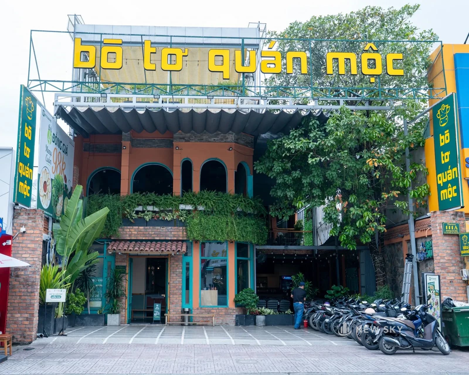 Mặt tiền Bò Tơ Quán Mộc: biển hiệu chữ vàng khổ lớn trên nóc, mặt nhà gạch đỏ phủ cây leo, cửa vòm sơn xanh.