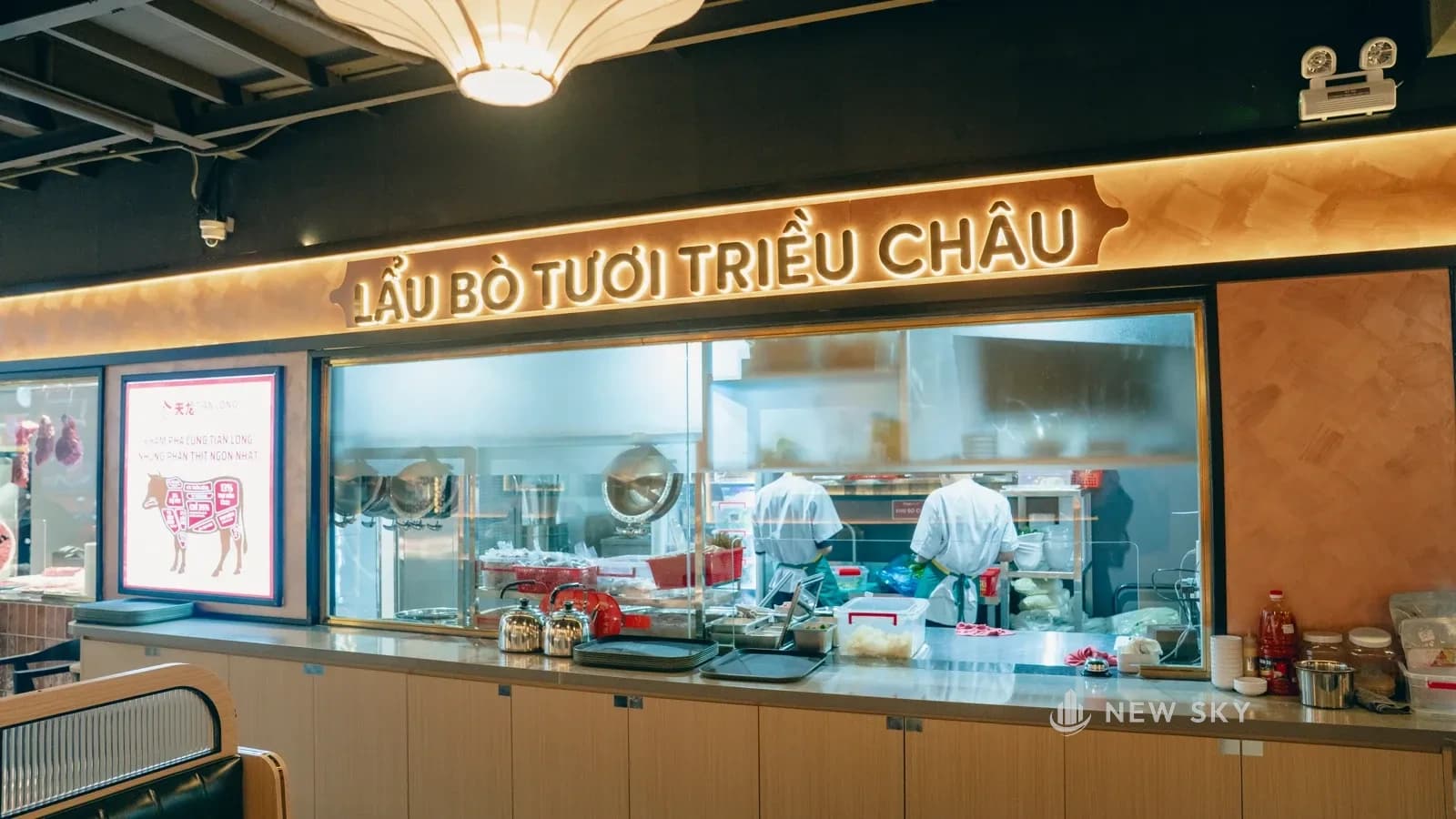 Khu bếp nhìn qua ô kính rộng, phía trên treo biển chữ nổi; mặt quầy lát đá sáng, thân tủ ốp gỗ.