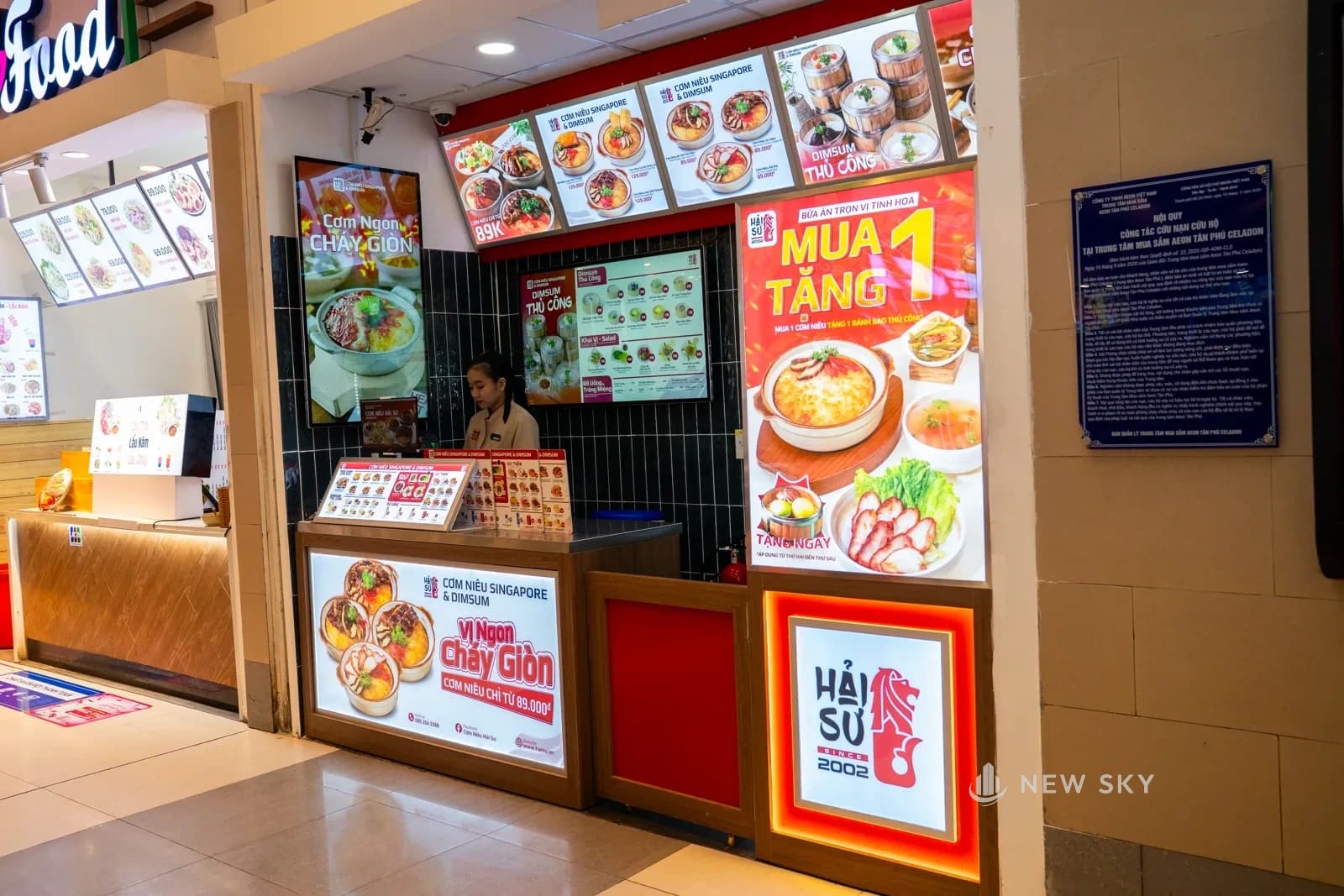 Mặt quầy nhìn nghiêng từ lối đi: tủ hộp đèn ảnh món chạy suốt thân quầy, tấm biển khuyến mại dựng cạnh mép quầy.