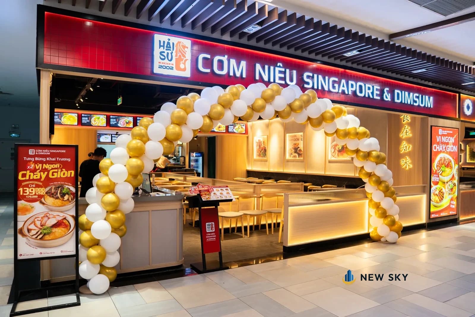 Mặt tiền Cơm Niêu Hải Sư tại Giga Mall nhìn từ hành lang tầng năm: biển hiệu đỏ chạy suốt bề ngang, cổng bóng bay dựng ngày khai trương, quầy đón khách ngay lối vào.