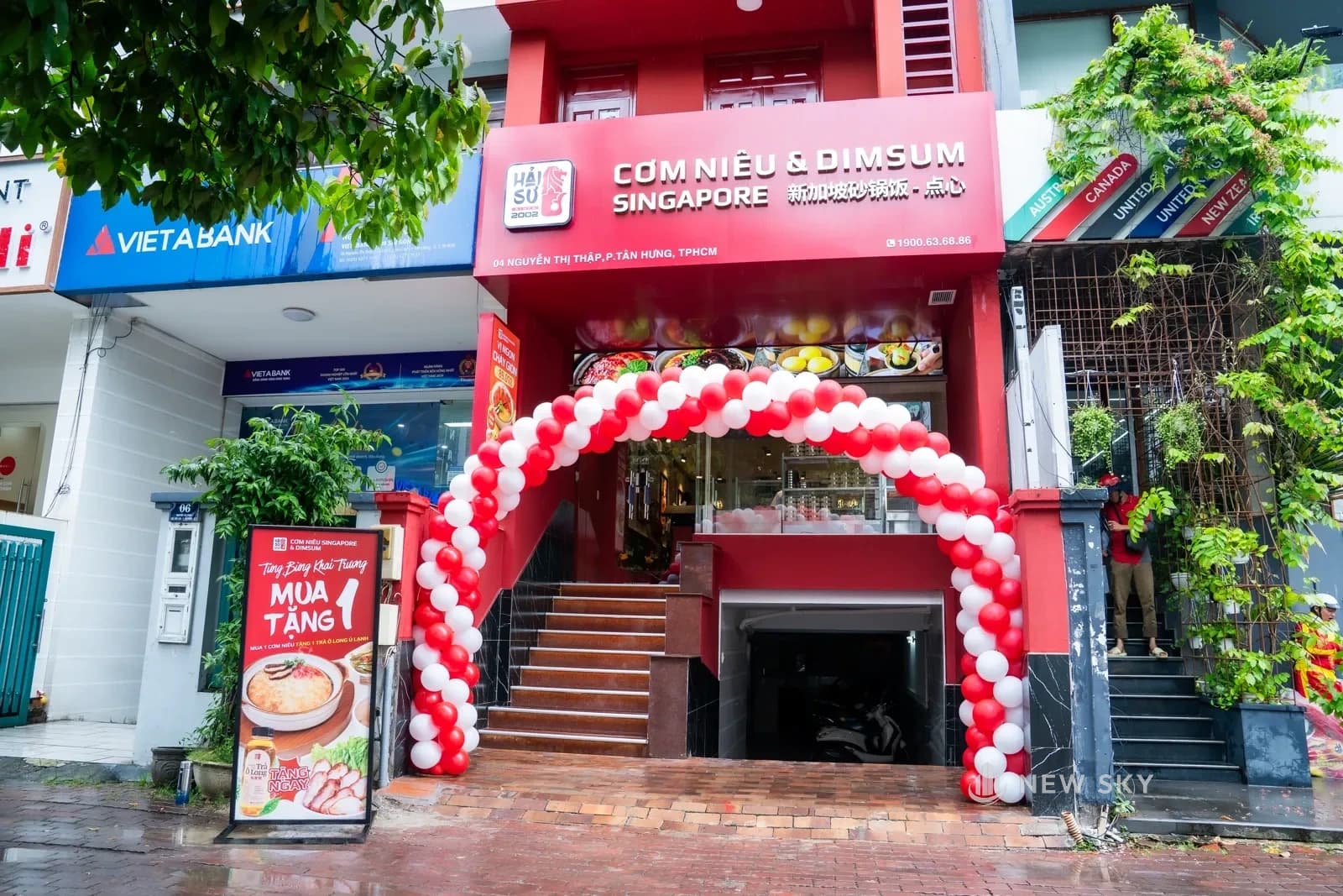 Mặt tiền nhà phố của Cơm Niêu Hải Sư trên đường Nguyễn Thị Thập: mặt nhà sơn đỏ, biển hiệu chạy suốt bề ngang, vế thang dẫn lên cửa kính, cổng bóng bay dựng ngày khai trương.