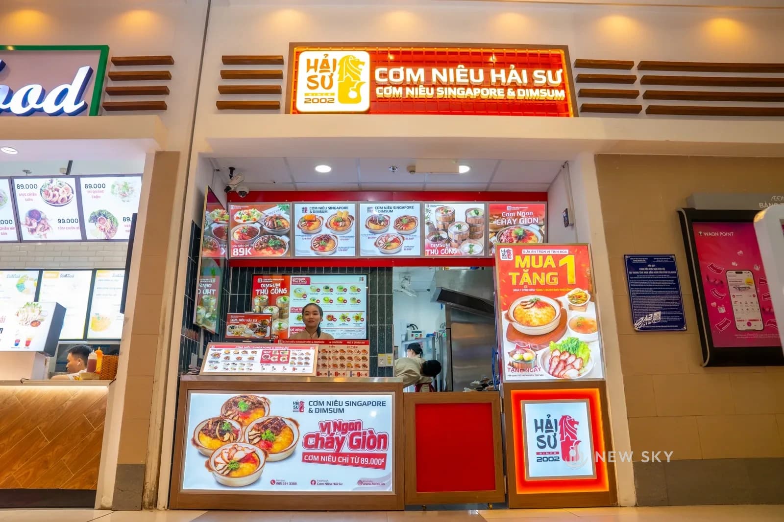 Quầy Cơm Niêu Hải Sư nhìn chính diện từ lối đi khu ẩm thực Aeon Mall Tân Phú Celadon: biển hộp đèn đỏ trên mảng tường ốp nan gỗ, dải hộp đèn ảnh món và hai màn hình thực đơn gắn trên vách gạch men xanh đen, nhân viên đứng sau quầy.