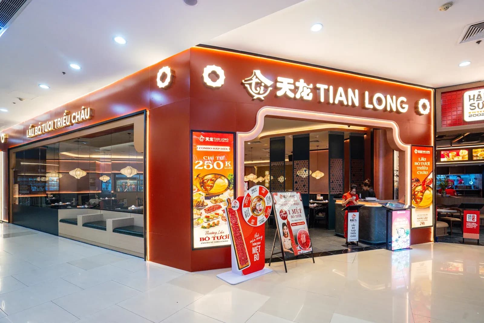 Mặt tiền Tian Long Vincom Bà Triệu nhìn chéo từ hành lang: cổng vòm đỏ cam ở góc, bên trái là vách kính dài có dãy ghế băng màu xanh sát chân kính, trên kính là biển "LẨU BÒ TƯƠI TRIỀU CHÂU"; bên phải là biển Hải Sư.